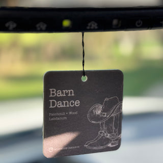 A rectangular car air freshener tag hanging from a mirror. The tag is dark gray with white text reading 'Barn Dance' and 'Patchouli + Wood Labdanum,' plus a white line drawing of a cowboy hat and boots.