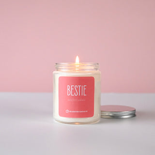 Lit candle in jar with a silver lid and  pink label reading 'BESTIE' on a light pink background