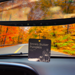 A car air freshener hanging from the rearview mirror inside a vehicle, with an autumn forest road visible through the windshield. The tag reads 'Brown Butter Pumpkin' and shows a pumpkin illustration.