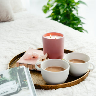 a blush colored ceramic candle with two cuips of tea on a ray placed on a bed 