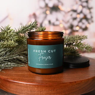 Candle jar with 'Fresh Cut Fraser' label on a wooden surface with Christmas decorations.