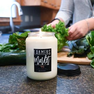 Candle labeled 'Farmers Market' on a kitchen counter with fresh produce.