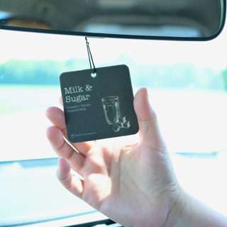 A hand holding a black car air freshener tag labeled 'Milk & Sugar' in front of a car window.
