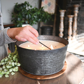 Person lighting a candle in a metal container on a wooden table with a plant in the background