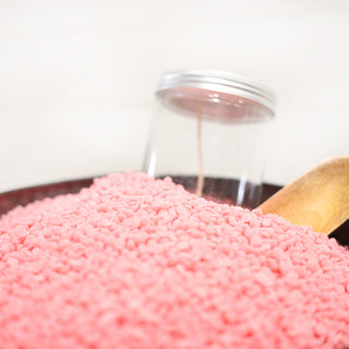 Pink wax beads in a metal bowl with a wooden spoon on a white background