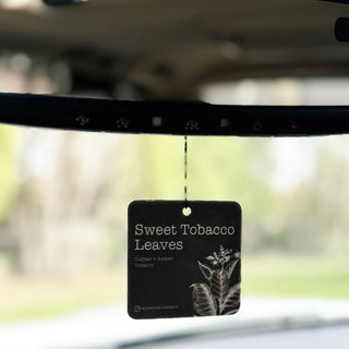 A square car air freshener hanging from a mirror inside a vehicle. The tag reads 'Sweet Tobacco Leaves' with a botanical leaf illustration on a blurred outdoor background.