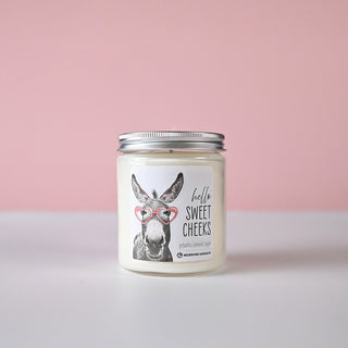 Clear candle jar with silver lid and a donkey illustration that says 'hello sweet cheeks' in black text against a pink background