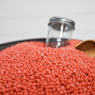 Red wax beads in a metal container with a glass jar and wooden scoop on a white background