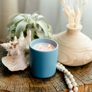 a cerelean ceramic candle on a stump with some beads, a shell, plant and a wood vase with flowers 