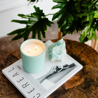 a tourquise ceramic lit candle next to a piece of  quartz on a cerial magazine on a stump next to a plant 