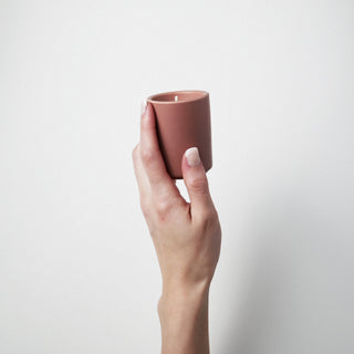 Candle in a blush colored container being held by a hand against a white background