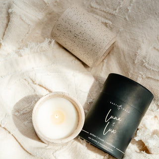 two speckled cream ceramic candle jars and a tube that goes with laying on a textured cream blanket 